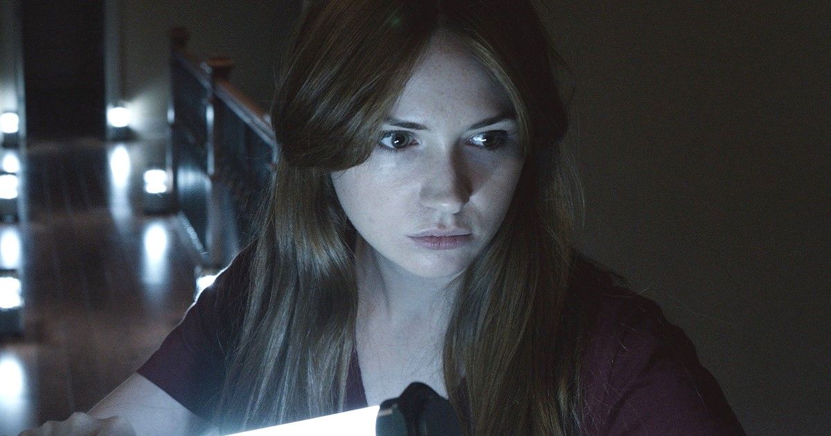 Karen Gillan looks off camera in a dim-lit room with a light in front of her. 