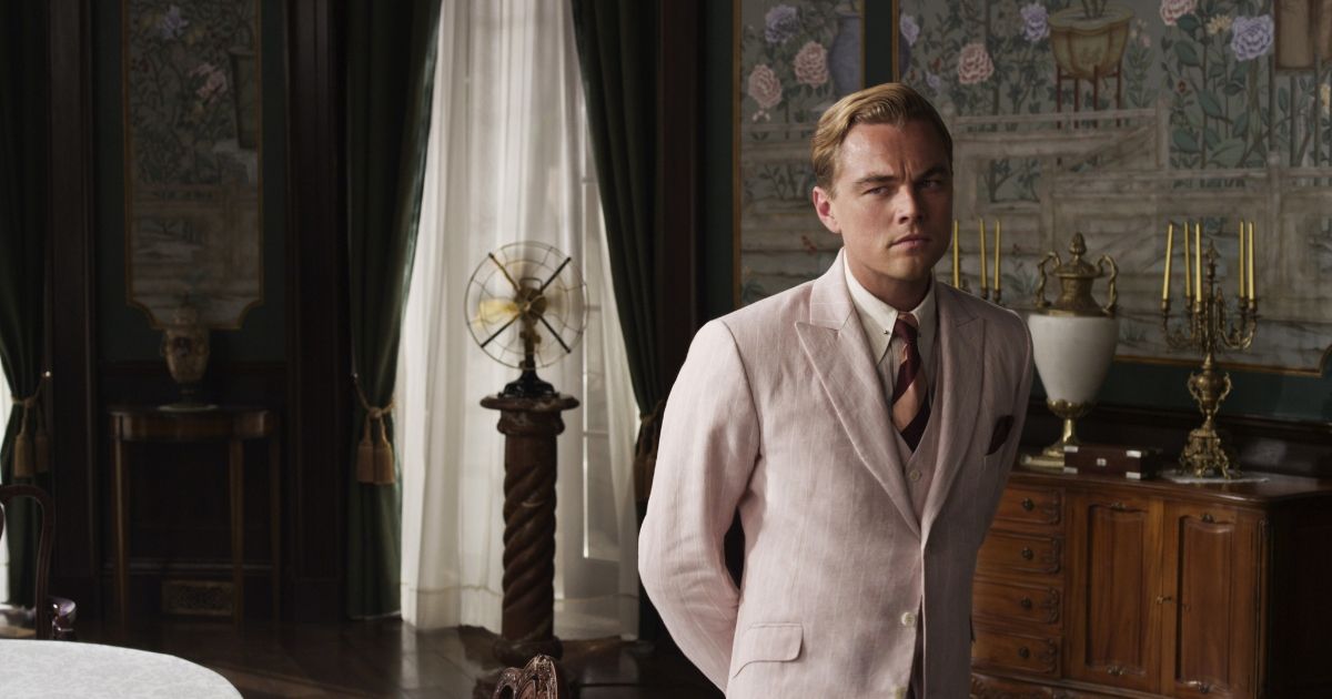 A man in a beige suits stands in the middle of an ornate room.