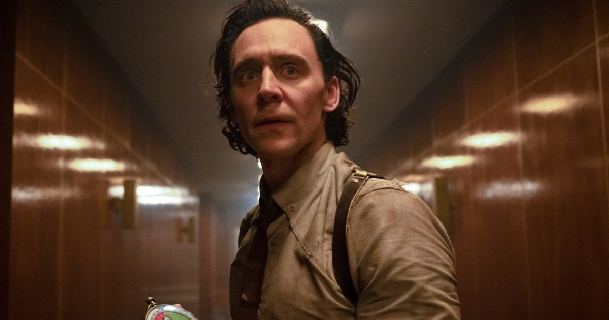 Loki holding a device in his hand, looking down a long hallway while wearing his tan collared shirt and suspenders in Loki.
