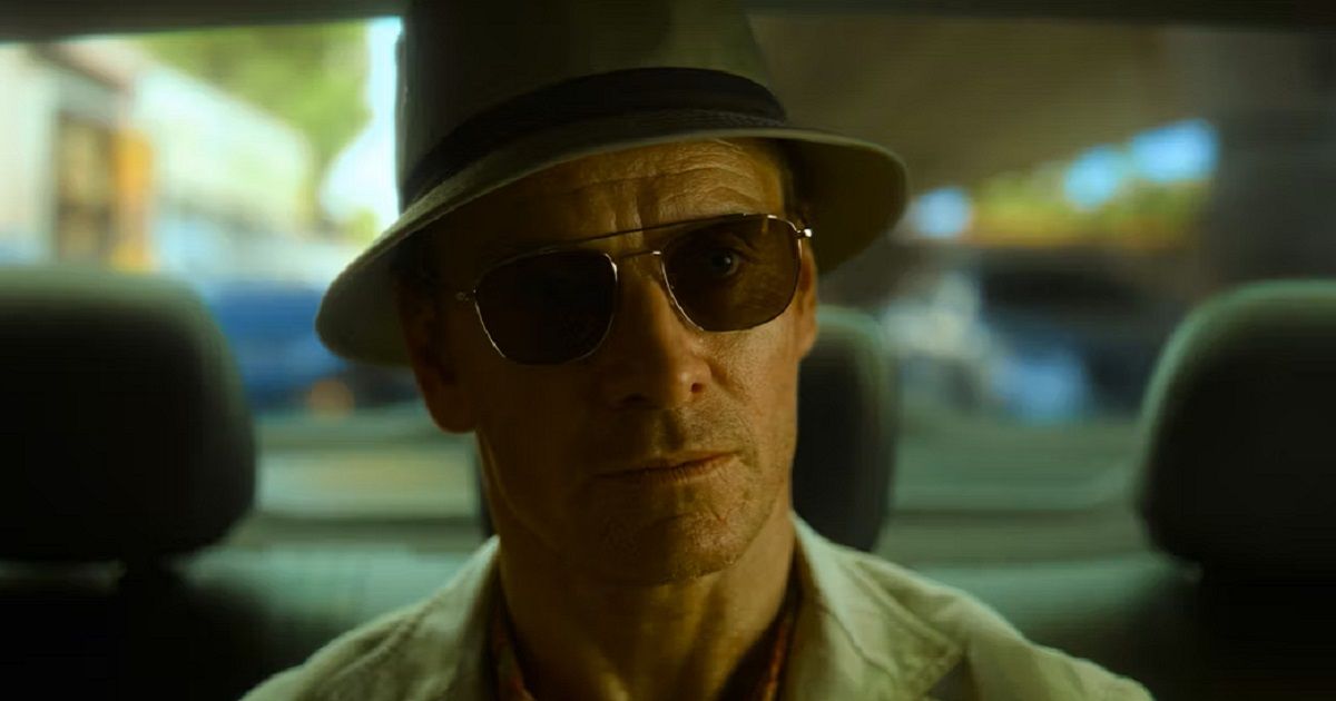 Michael Fassbender usando chapéu, óculos e jaqueta leve bege sentado no banco de trás de um carro em The Killer.