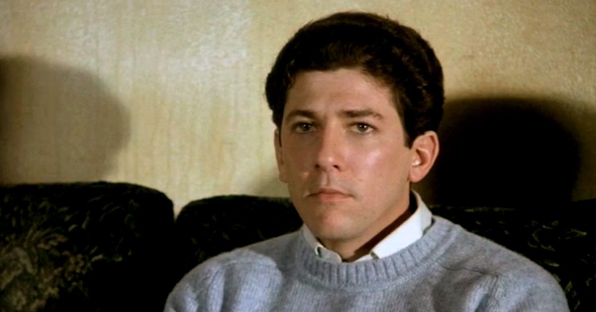 Peter Riegert sitting on a couch with a sweater and collared shirt in Animal House