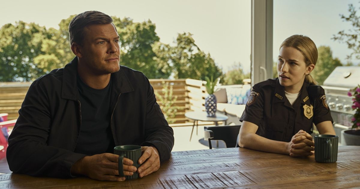Alan Ritchson as Jack Reacher drinking a cup of coffee at a table outside with a police officer next to him in Reacher.
