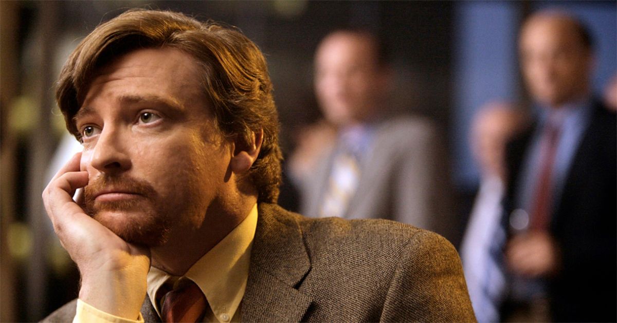 Rhys Darby in a brown suit and yellow shirt with two men standing behind him out of focus in Flight of the Conchords.