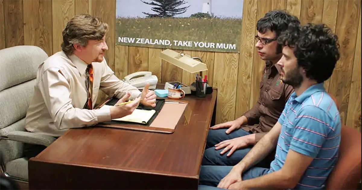 Rhys Darby sentado em sua mesa com Bret McKenzie e Jemaine Clement em Flight of the Conchords