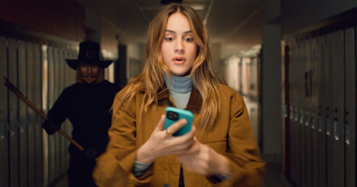 Nell Verlaque as Jessica wearing a turtle neck and brown jacket, with John Carver approaching behind carrying an axe in the high school locker room in Thanksgiving (2023)