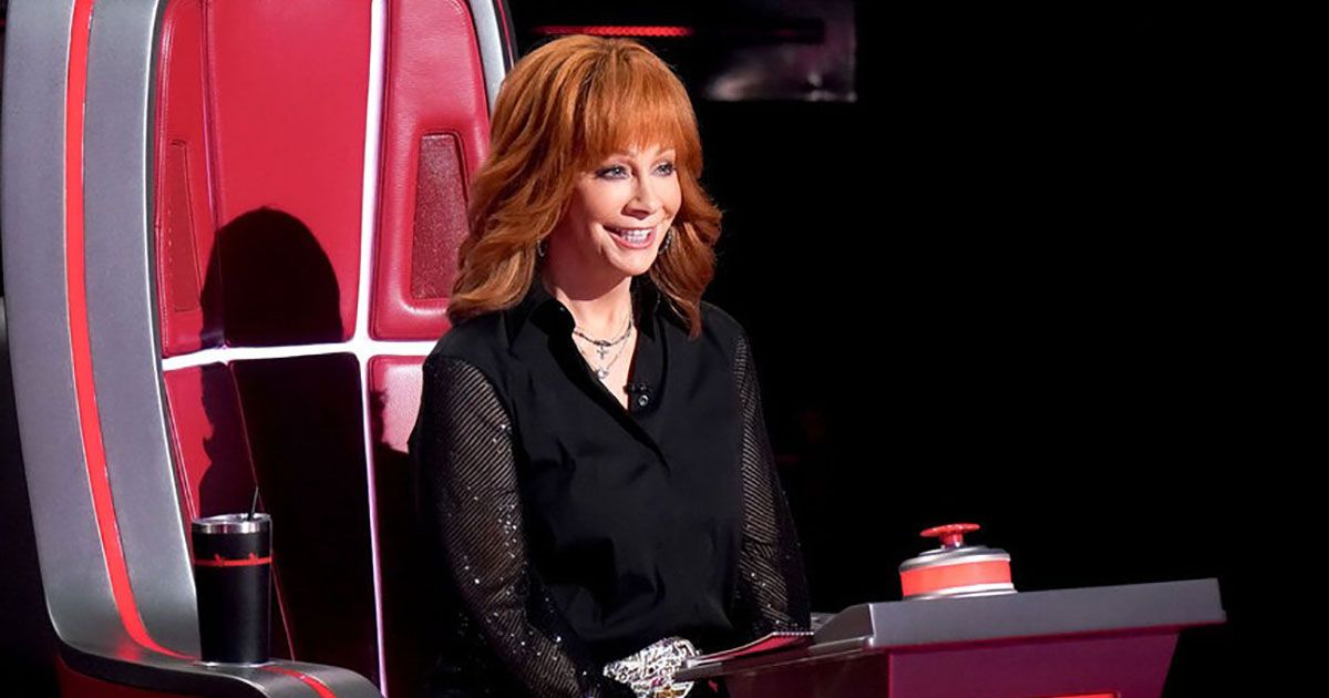 Reba McIntire sentada em uma cadeira, campainha na frente dela e sorrindo em cena do The Voice.