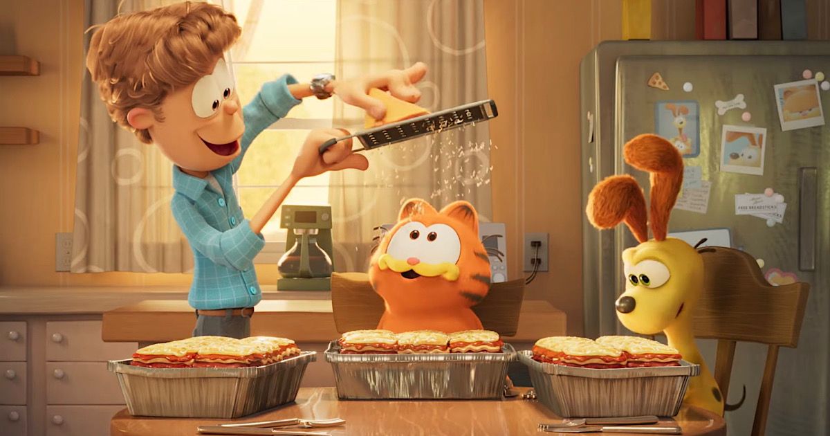 A man grating cheese over trays of lasagna in a kitchen as Chris Pratt's Garfield looks with amazement, and another dog sits on a chair preparing to eat in Garfield.
