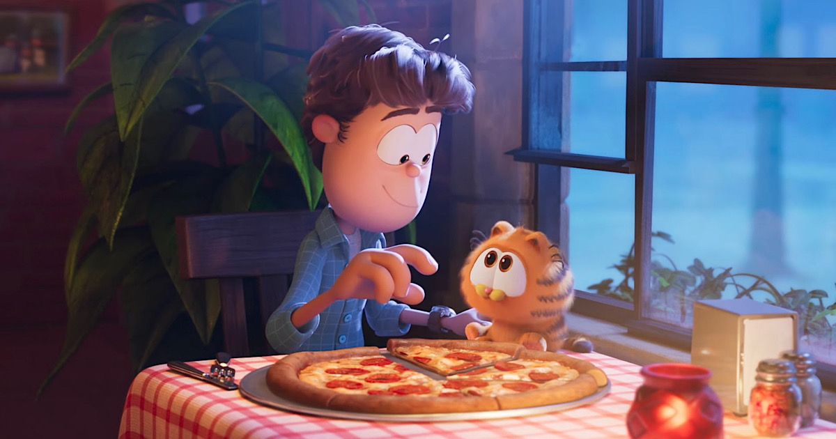 Jon and Chris Pratt's Garfield as a kitten sits in a restaurant with a large pizza on the table as Garfield looks interested in the food and Jon prepares to feed him in Garfield