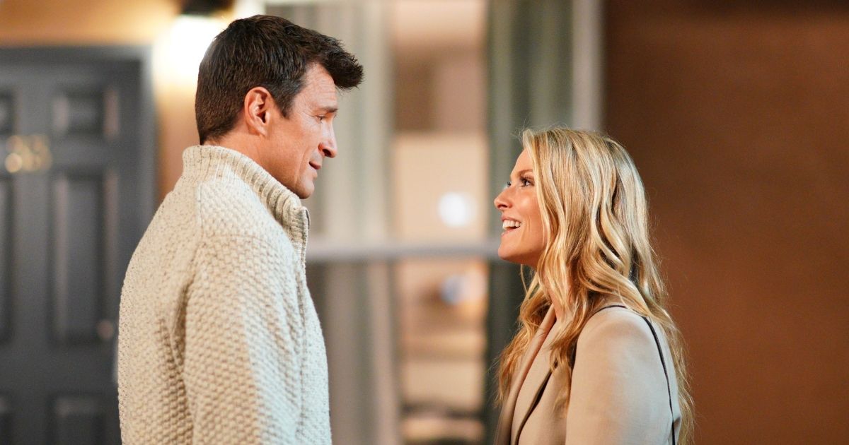 Nathan Fillion as John Nolan, wearing a light colored sweater, and Ali Larter as Grace Sawyer, wearing a tan jacket and black purse as the two talk in front of a house in The Rookie.