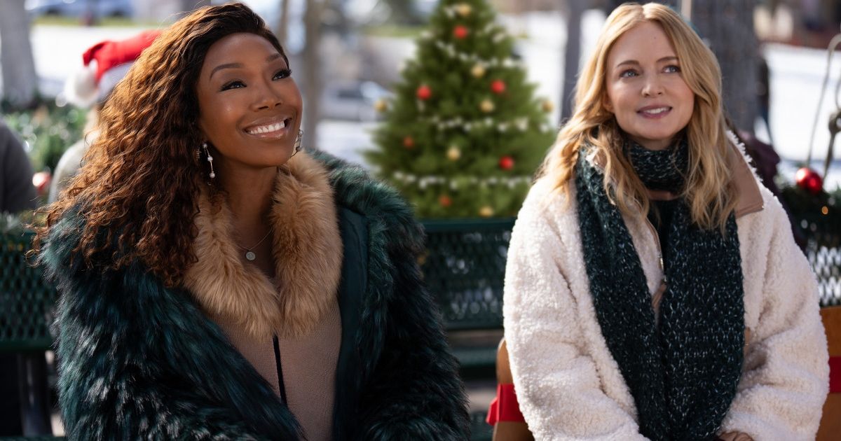 Heather Graham as Charlotte Sanders, wearing a white coat and large scarf, smiling and looking at something off camera with Brandy Norwood as Jackie Jennings, wearing a green fur coat and tan undercoat, smiling with a Christmas tree in the background in Best. Christmas. Ever! 