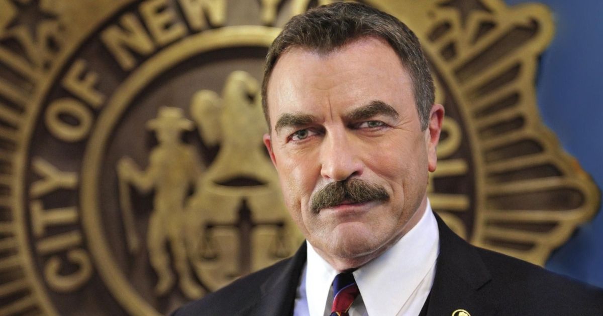 Tom Selleck as Frank Reagan in Blue Bloods, wearing a suit, standing in front of a large seal for the City of New York in Blue Bloods.