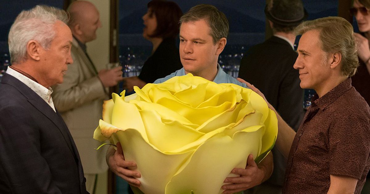 Udo Kier as Konrad, Matt Damon as Paul Safranek, and Christoph Waltz as Dusan Mirkovic with Damon holding a large yellow flower in his hands at a party in Downsizing.