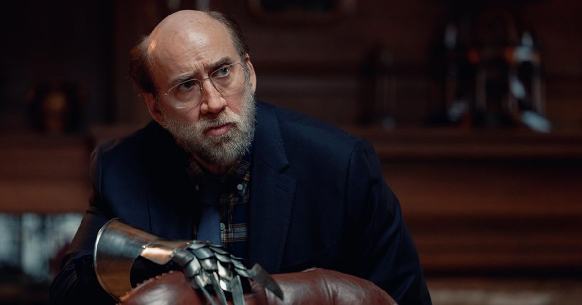 Nicolas Cage in a blue suit jacket, wearing an armored glove on one hand clenching a leather chair in Dream Scenario