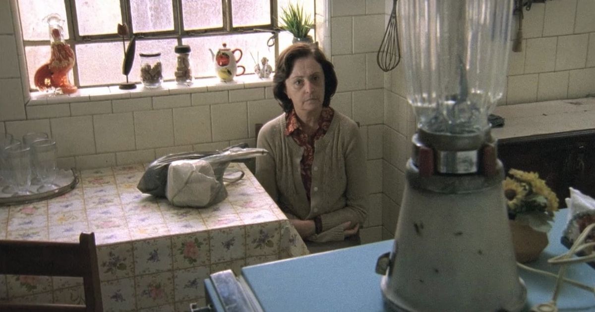 Uma senhora idosa sentada à mesa da cozinha olha para o liquidificador, aparentemente absorta em sua presença.