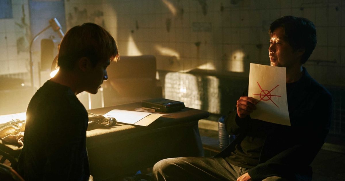 Two men sit in a dimly lit police precinct, with one of them holding up a piece of paper with a symbol on it.