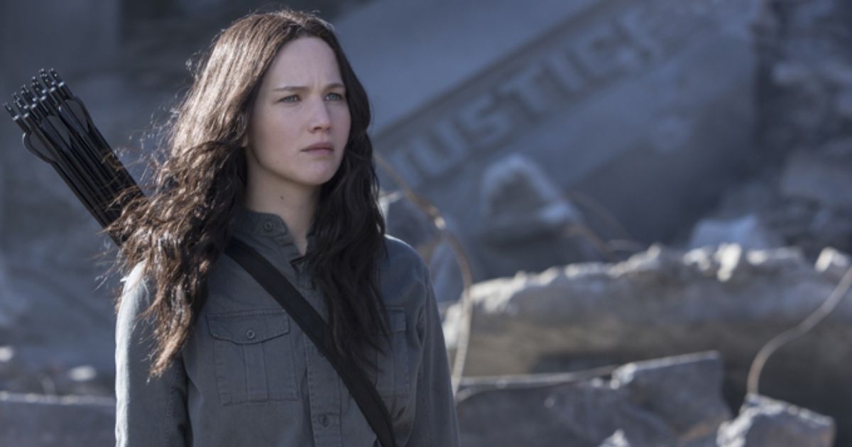 Jennifer Lawrence as Katniss Everdeen in a grey button up shirt with a quiver full of arrows on her back, looking at destroyed rubble in The Hunger Games: Mockingjay