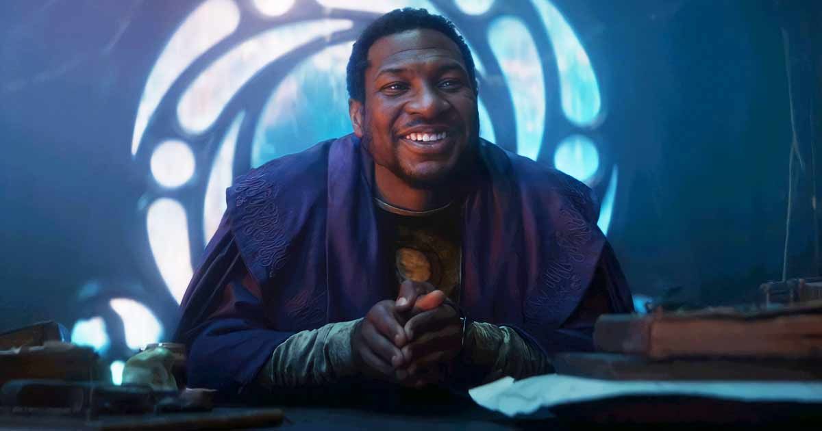 Jonathan Majors as He Who Remains in Loki, wearing his purple robes sitting at his desk with a glowing window behind him smiling at Sylvie and Loki.