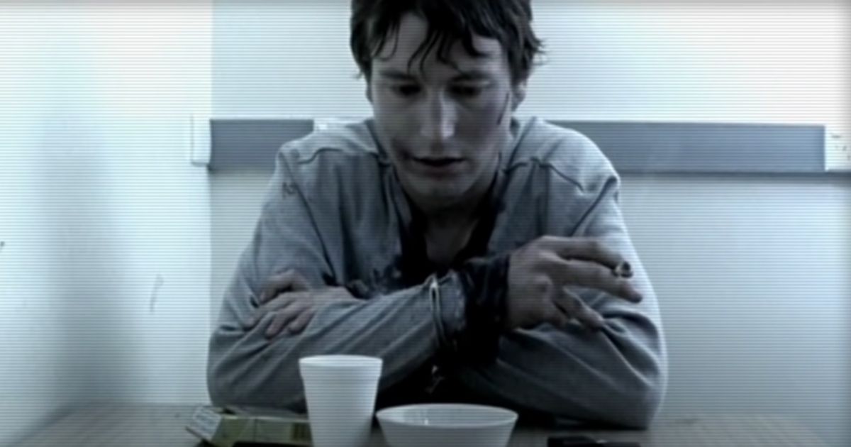 Leigh Whannell as David in Saw 0.5, chained to a desk, smoking a cigarette with a bowl and cup in front of him on the desk.