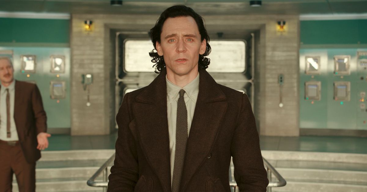 Tom Hiddleston as Loki, wearing his brown coat and white collared shirt in the TVA, with Owen Wilson's Mobius in a similar outfit in the background.