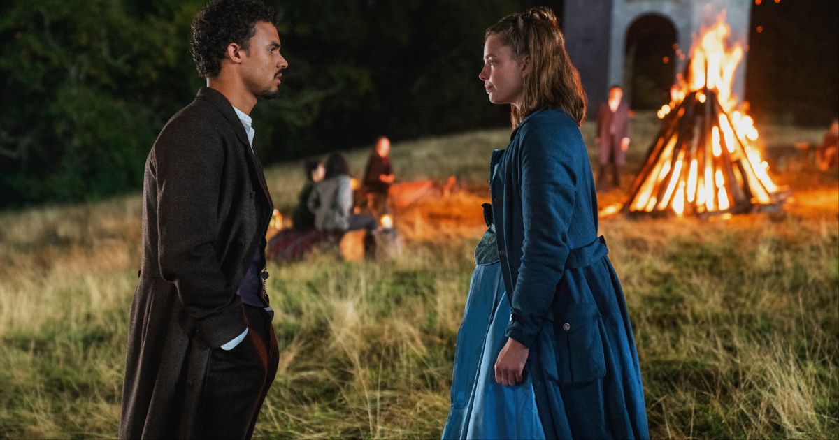 Matthew Broome as Guy win a long coat and nice pants, and Kristine Froseth as Nan wearing a blue dress with a large bonfire behind them in The Buccaneers