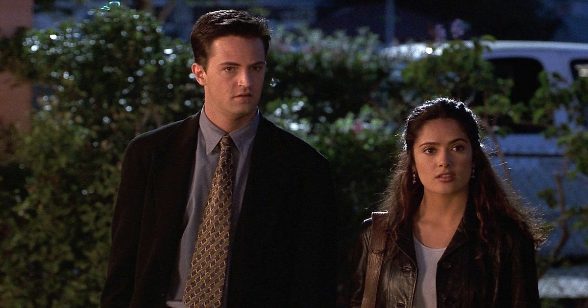 Matthew Perry wearing a black suit jacket, blue shirt, and brown and gold tie and Salma Hayek standing next to him in a black jacket with her purse in Fools Rush In