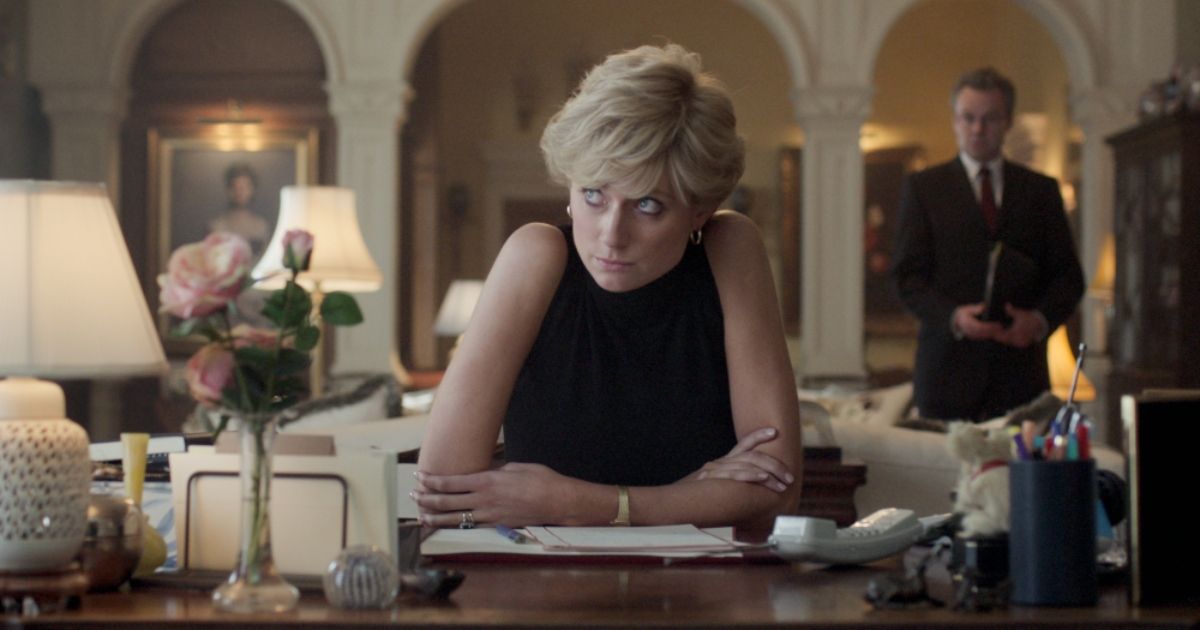 Elizabeth Debicki as Princess Diana wearing a black dress sitting at a desk with papers on it and a man in a suit behind her in The Crown.