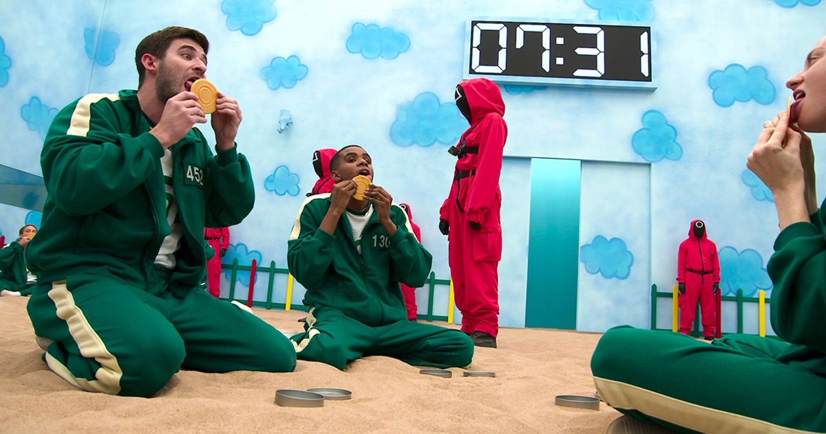 A group of participants wearing green and white clothes look cookies while sitting in sand in Squid Game: The Challenge