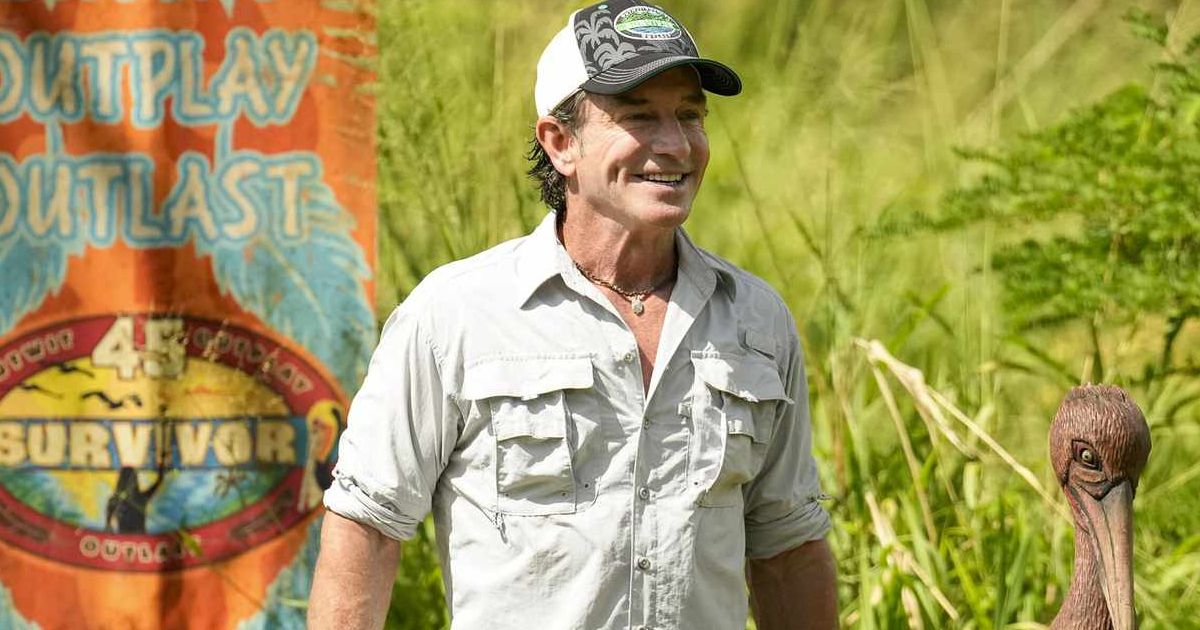 Jeff Probst in Survivor Season 45, wearing a light-colored collared shirt with his sleeves rolled up, next to a Survivor flag and immunity idol, standing in the jungle of Fiji.