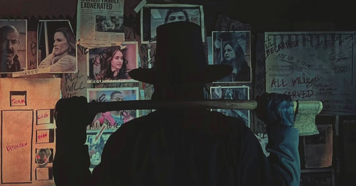 A man dressed in all black with a pilgrims hat on and axe resting over his shoulders, with a bunch of photos of victims hung up on his wall in Thanksgiving.