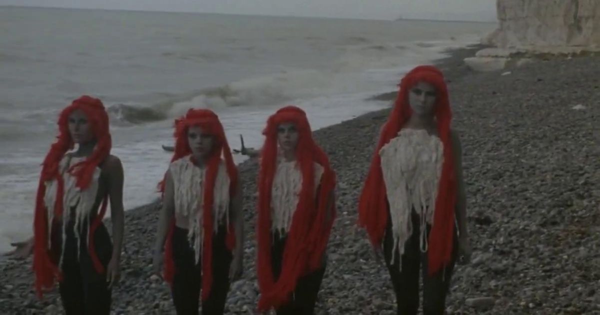 Quatro mulheres estranhamente vestidas estão em uma praia pedregosa.