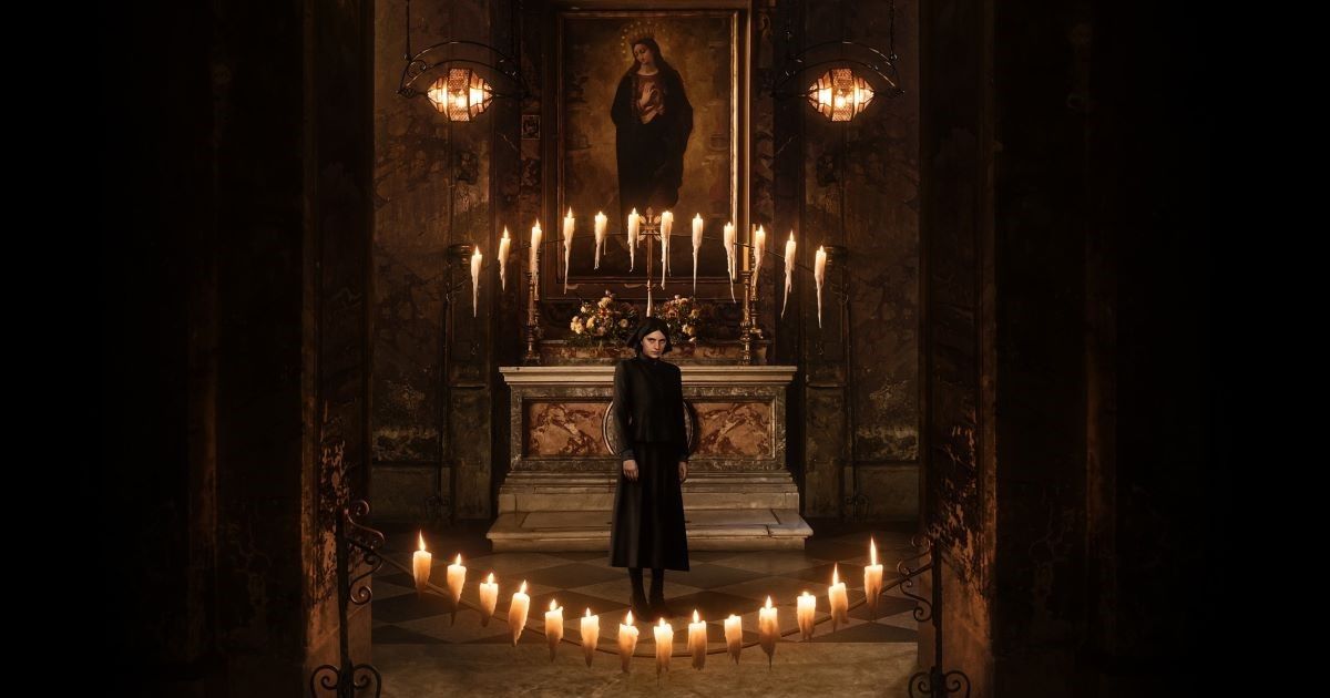 Nell Tiger Free in The First Omen, with candles and the Virgin Mary painting on the wall, with Free dressed in all black looking at the camera from a distance in The First Omen.