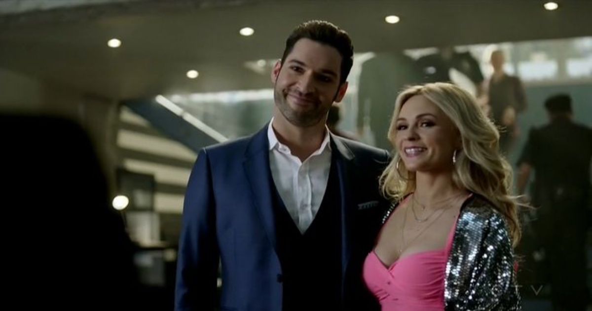 Lucifer wears a navy suit as he poses for a photo with a blonde woman in a pink top