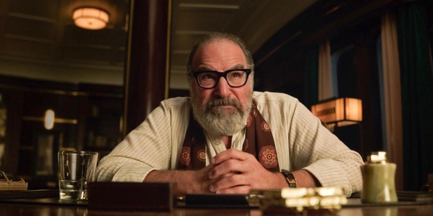 Mandy Patinkin as Rufus Cotesworth sitting at a desk staring at someone across from him in Death and Other Details