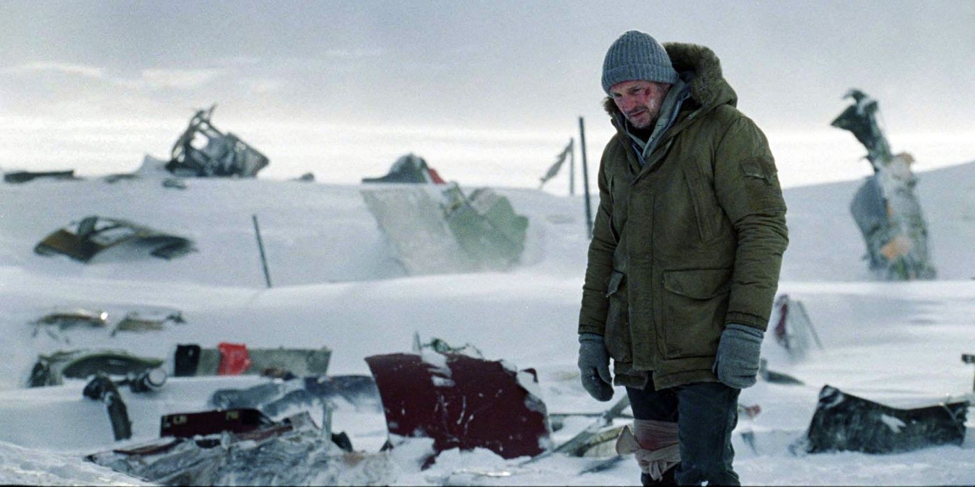 An injured man in a coat and toque stands in the middle of a plane crash wreckage.