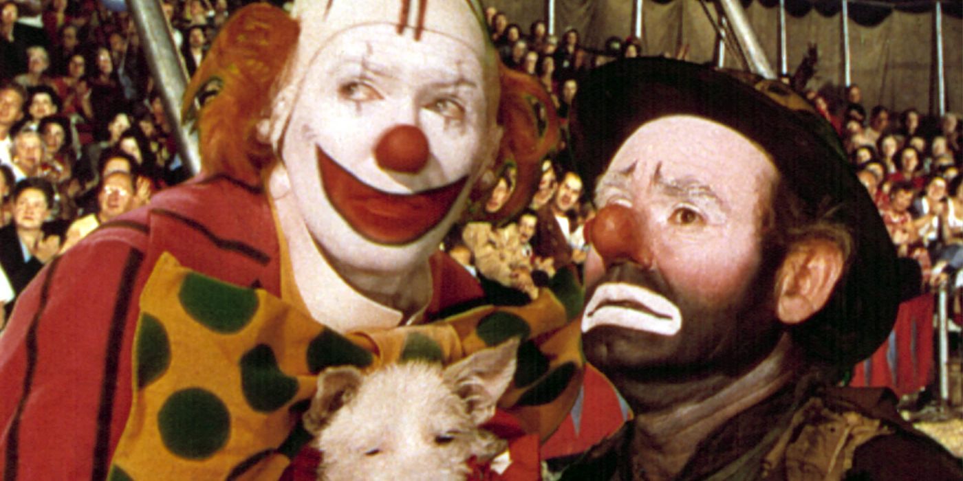 Two clowns, one in happy make-up and the other in sad, stand in a circus tent surrounded by an audience