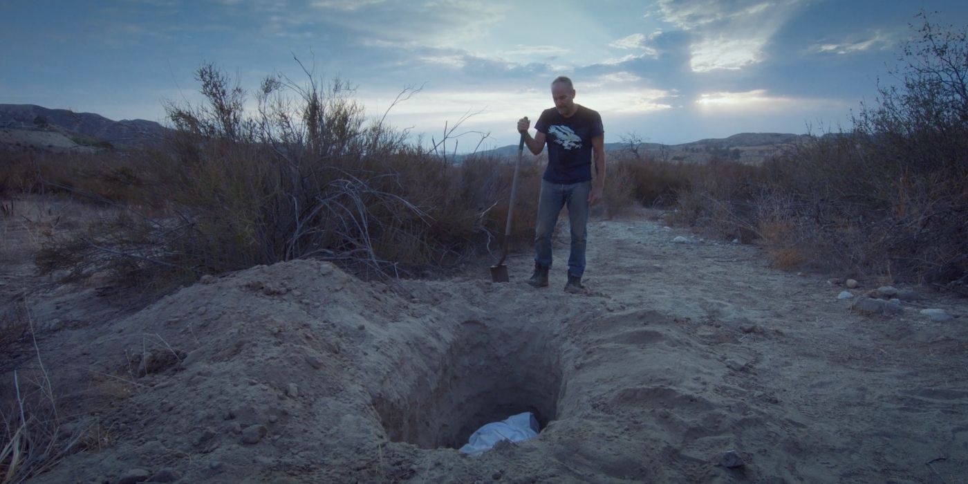Clint Jordan as Dwayne, standing beside a freshly dug grave in the middle of a desert and holding a shovel, in Bliss