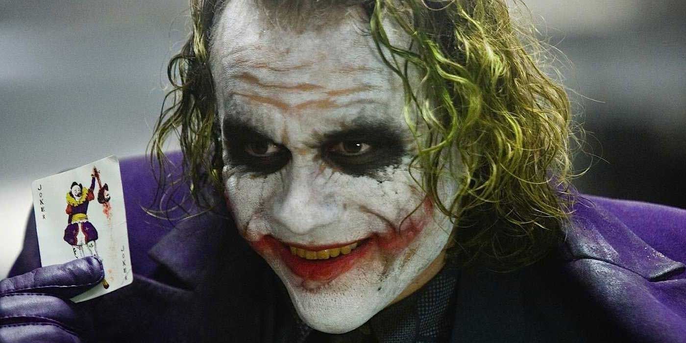 Heath Ledger as The Joker, grinning as he holds a joker playing card, in The Dark Knight