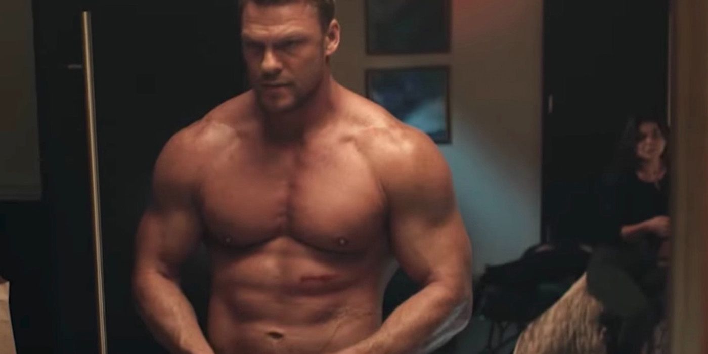 Alan Ritchson as Jack Reacher without a shirt with a cut on his abdomen and a woman out of focus behind him in Reacher 