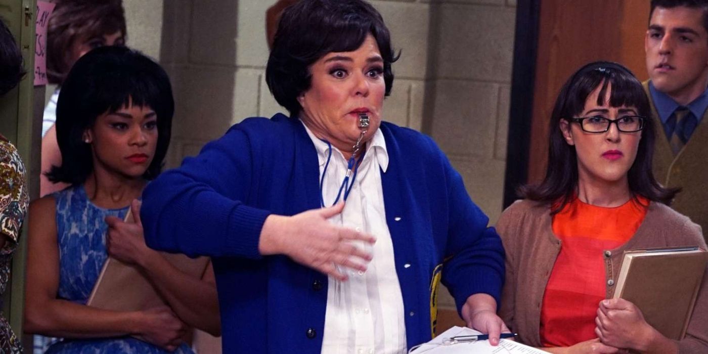 Rosie O'Donnell wearing a blue cardigan and blowing a whistle as the gym teacher on Hairspray Live