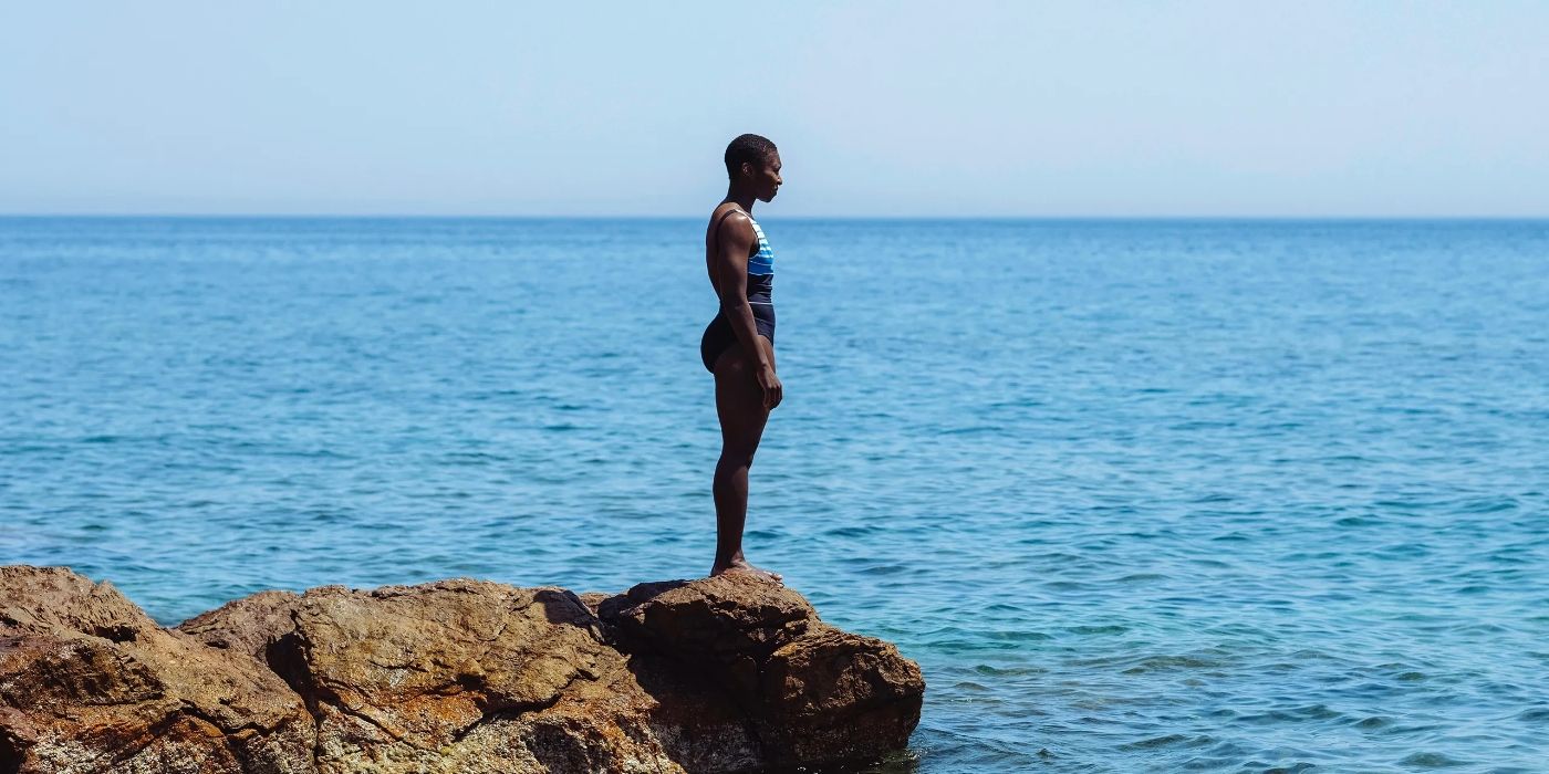 Cynthia Erivo as Jacqueline, wearing a swimsuit and standing at the edge of a rock by the sea