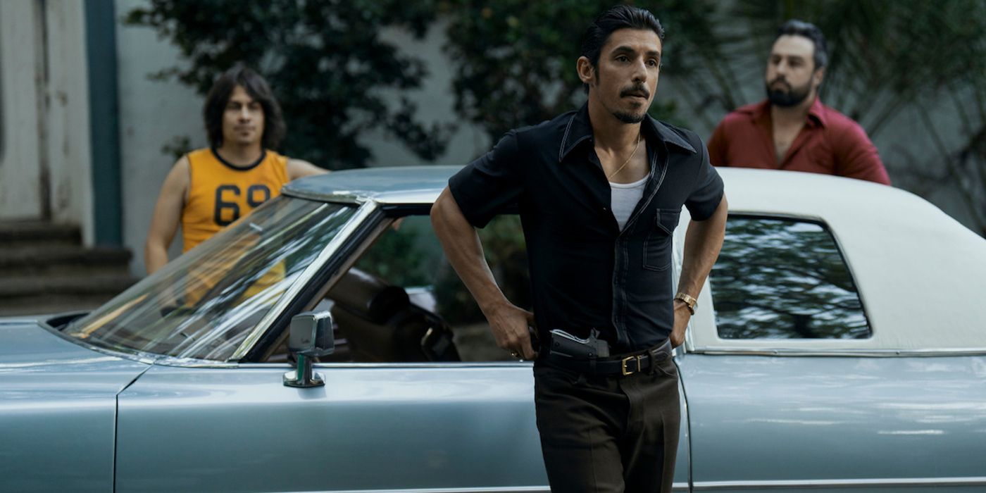 Alberto Guerra as Dario stands by a car wearng a black shirt with a gun with two men behind him in Griselda