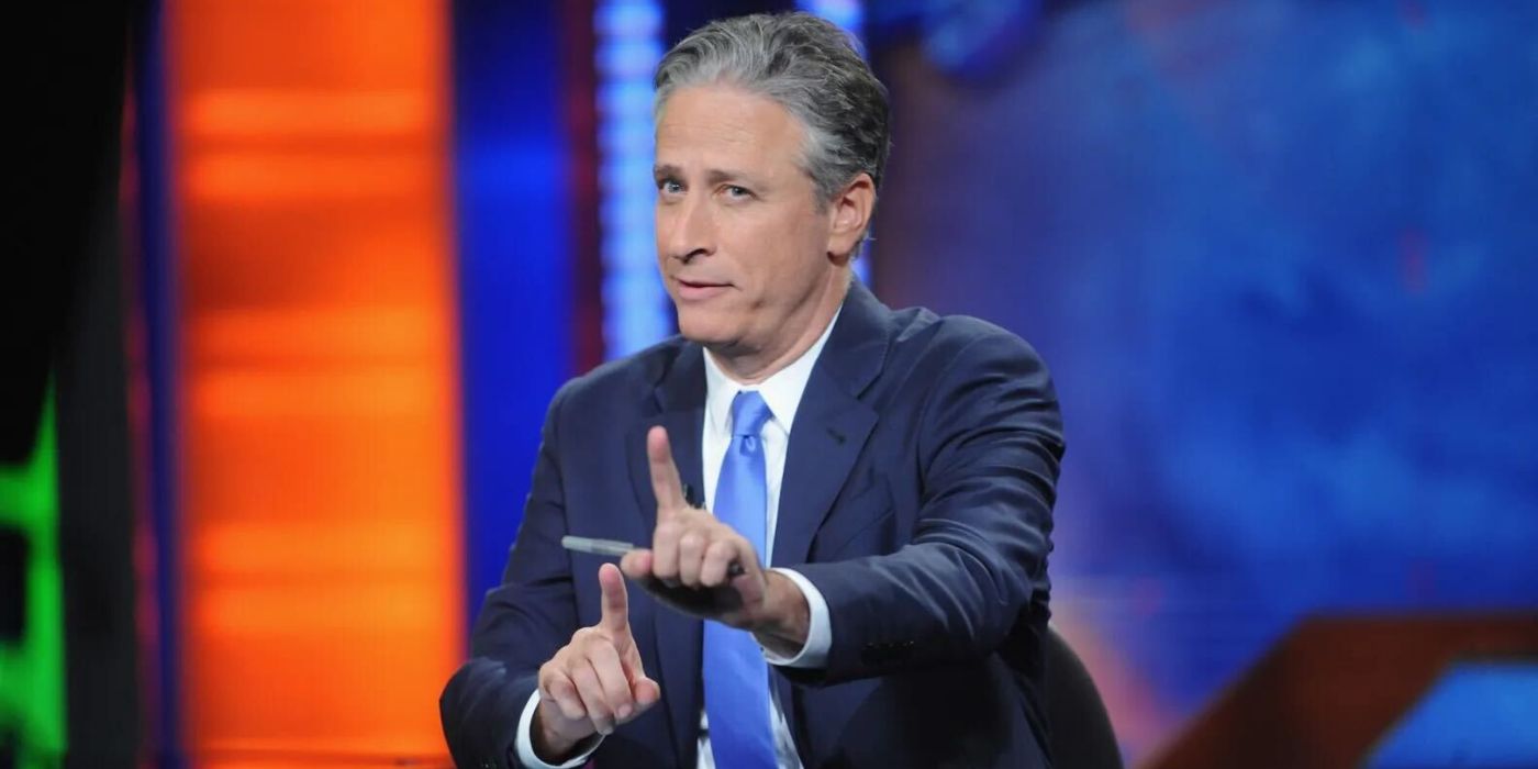 Jon Stewart wearing a blue suit and tie holding his finger up at the camera on The Daily Show