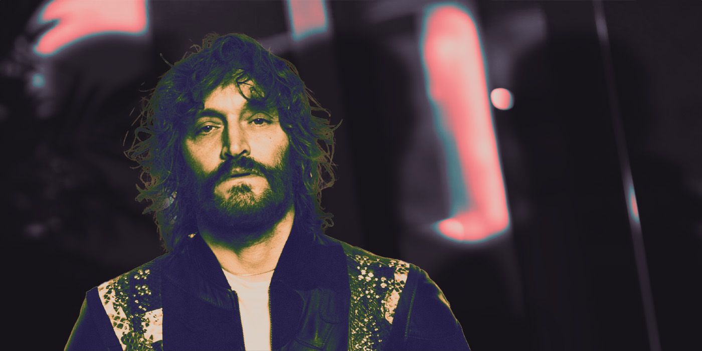 Vincent Gallo looking at the camera with long hair and a beard with silhouettes behind him