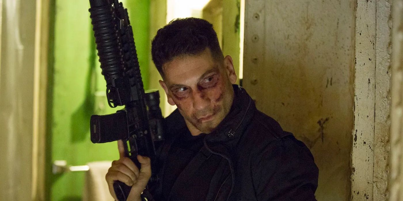 Jon Bernthal as the Punisher wearing an all black outfit carrying a large assault rifle in Punisher