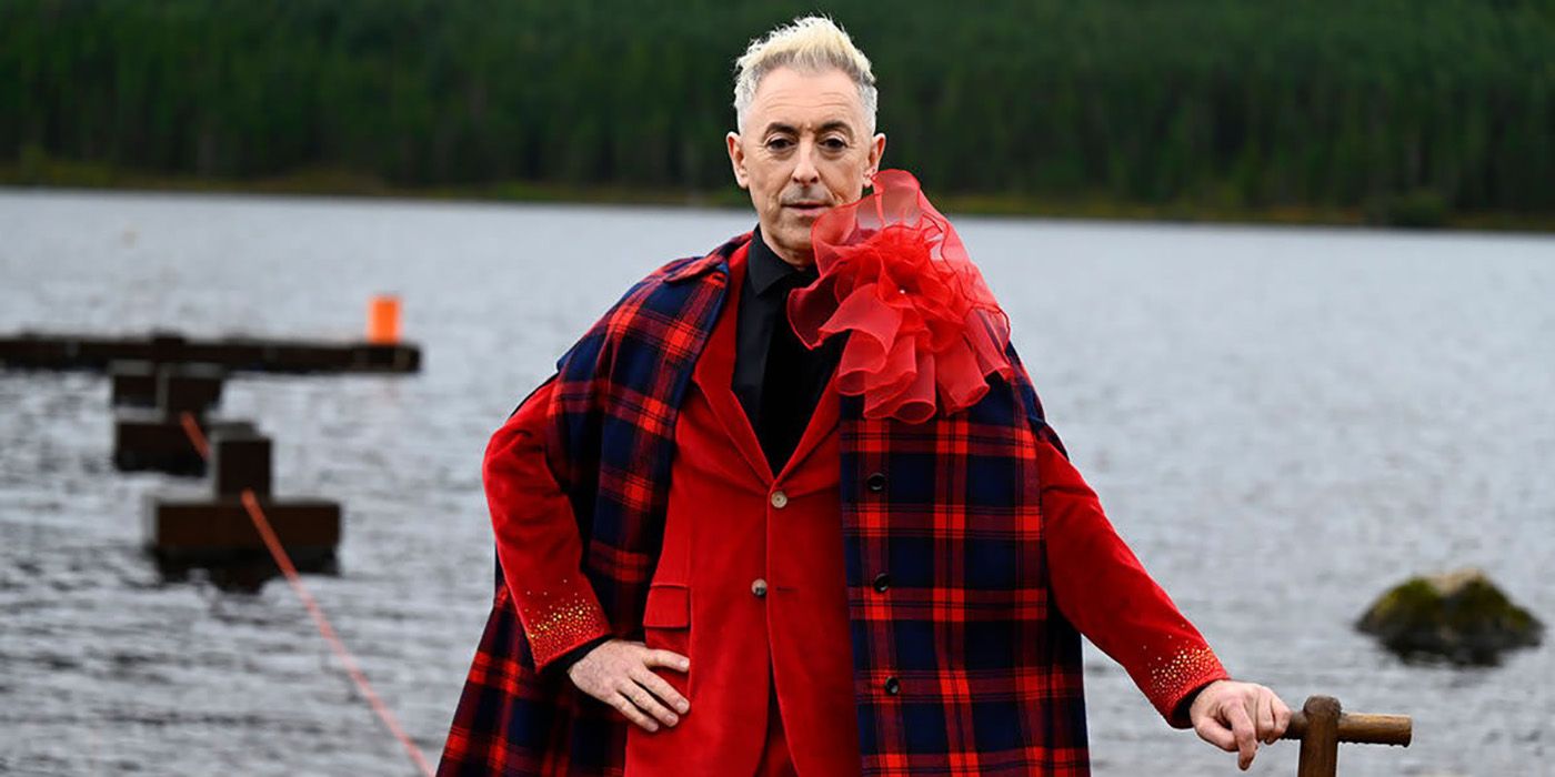 Alan Cummings dressed in red tartan standing by the water in a scene from The Traitors.