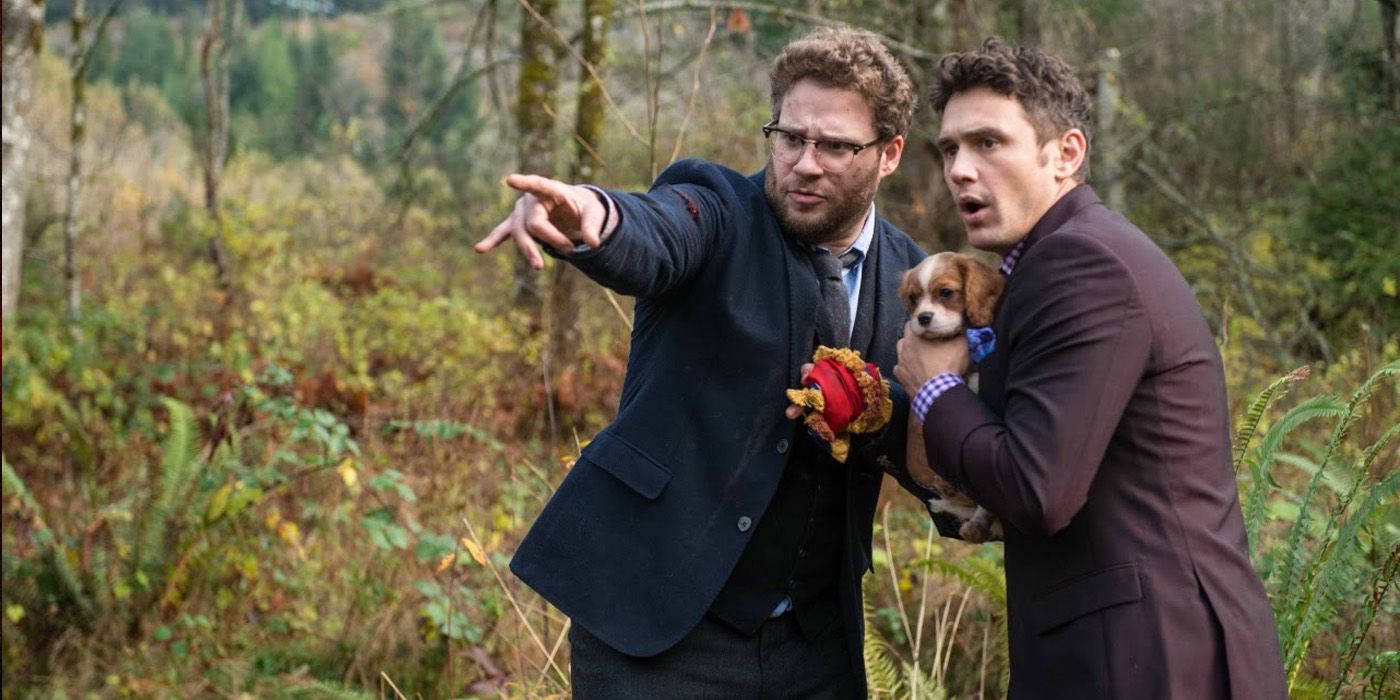 James Franco and Seth Rogen wearing suits holding a puppy in the woods pointing off-screen in The Interview