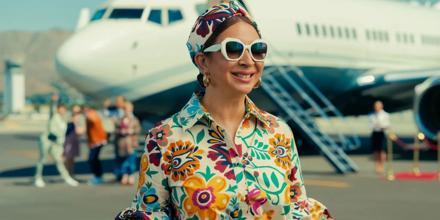 Maya Rudolph wearing a colorful shirt and hat standing in front of a private jet in Loot