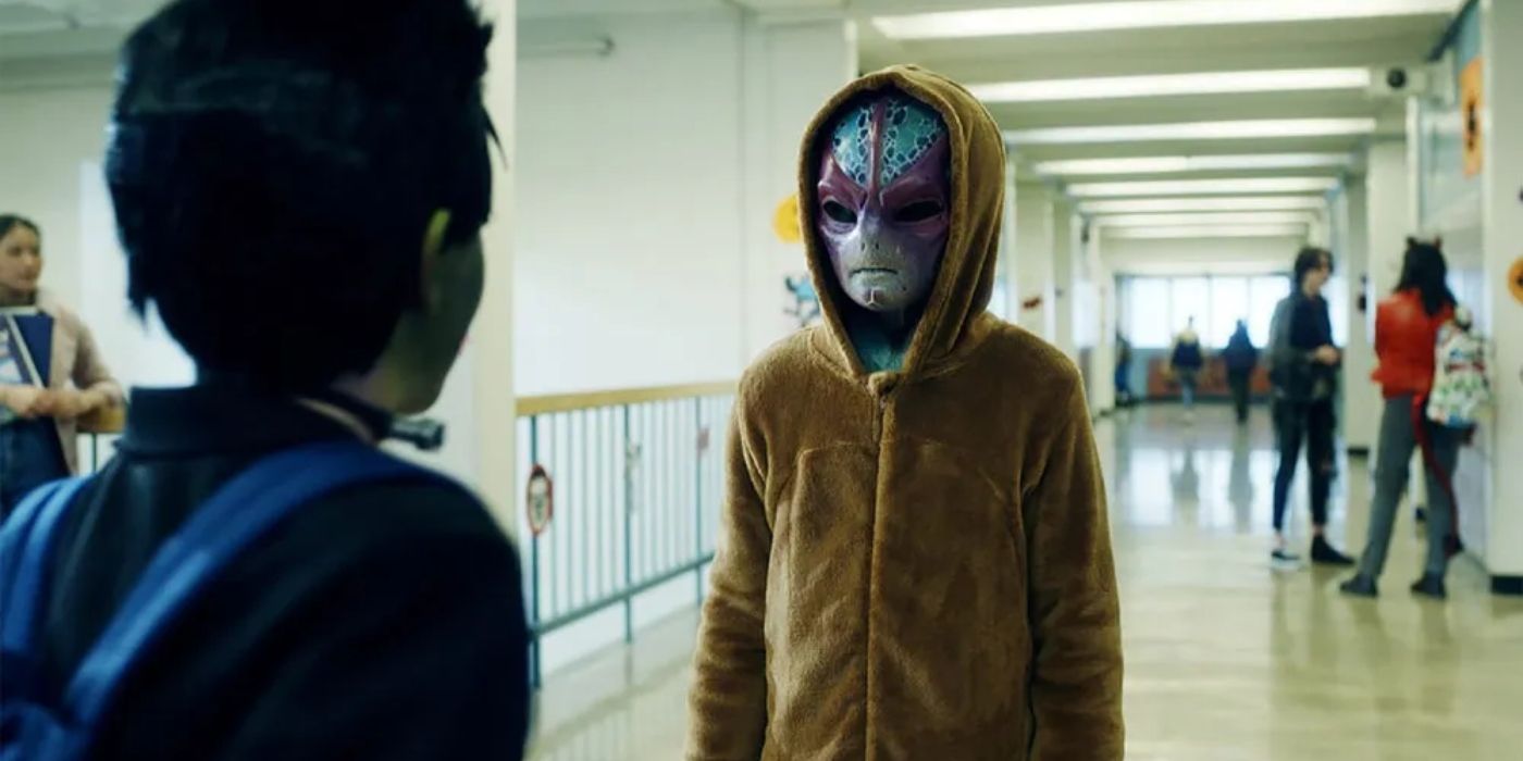 Alan Tudyk as Harry in his alien form wearing a brown hoodie standing in a school hallway in Resident Alien