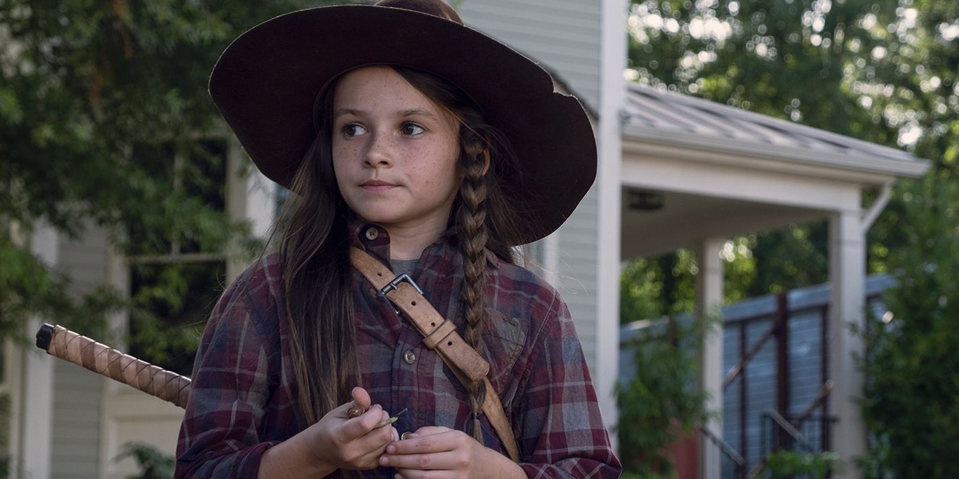 Judith standing with a police hat on next to houses in Alexandria in The Walking Dead.