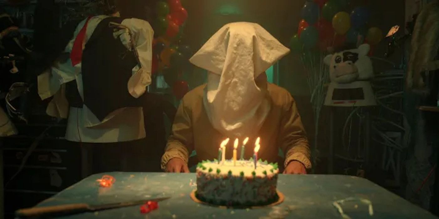A person with a sack over their head sitting in front of a birthday cake.